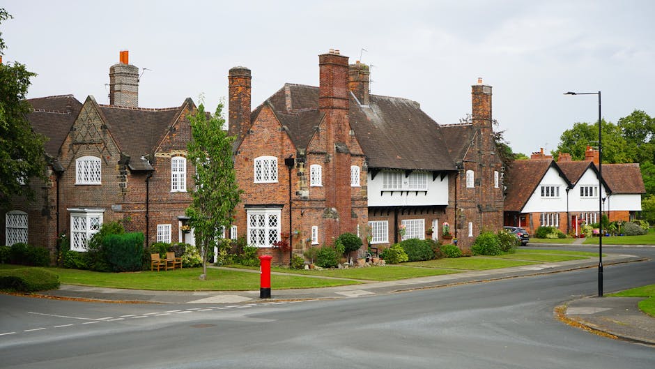 A row of traditional brick residential buildings with pitched roofs and multiple chimneys, situated along a quiet residential street in Swiss Cottage, Finchley Road. The houses feature white-framed windows, some with decorative leaded glass, and small front gardens with maintained shrubs and trees. In the foreground, the paved street includes a red post box near the curb, black street lamps, and a pedestrian sidewalk. The scene is well-lit with natural daylight, illustrating a peaceful neighbourhood environment that could be typical for home relocation or furniture transport services provided by Movers Swiss Cottage. This image captures the exterior setting where moving activities such as loading furniture onto vans or packing are likely to take place, highlighting the importance of professional removals in this area.