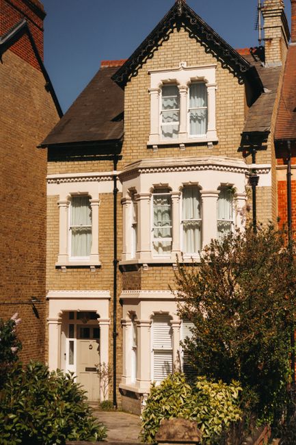 A row of traditional brick residential buildings with pitched roofs and multiple chimneys, situated along a quiet residential street in Swiss Cottage, Finchley Road. The houses feature white-framed windows, some with decorative leaded glass, and small front gardens with maintained shrubs and trees. In the foreground, the paved street includes a red post box near the curb, black street lamps, and a pedestrian sidewalk. The scene is well-lit with natural daylight, illustrating a peaceful neighbourhood environment that could be typical for home relocation or furniture transport services provided by Movers Swiss Cottage. This image captures the exterior setting where moving activities such as loading furniture onto vans or packing are likely to take place, highlighting the importance of professional removals in this area.