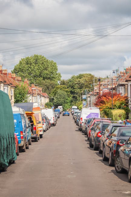 A white delivery van belonging to Movers Swiss Cottage is parked on a narrow, inclined street in Swiss Cottage, with its front facing uphill. The van is positioned close to the side of a multi-storey,